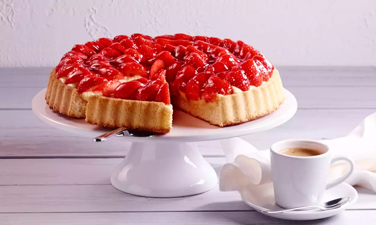 Mansikkalohkoilla peitetty torttu koristeltuna kiiltävällä punaisella kiilteellä, keltainen pohja korkeareunainen ja tarjottimella valkoisella jalustalla