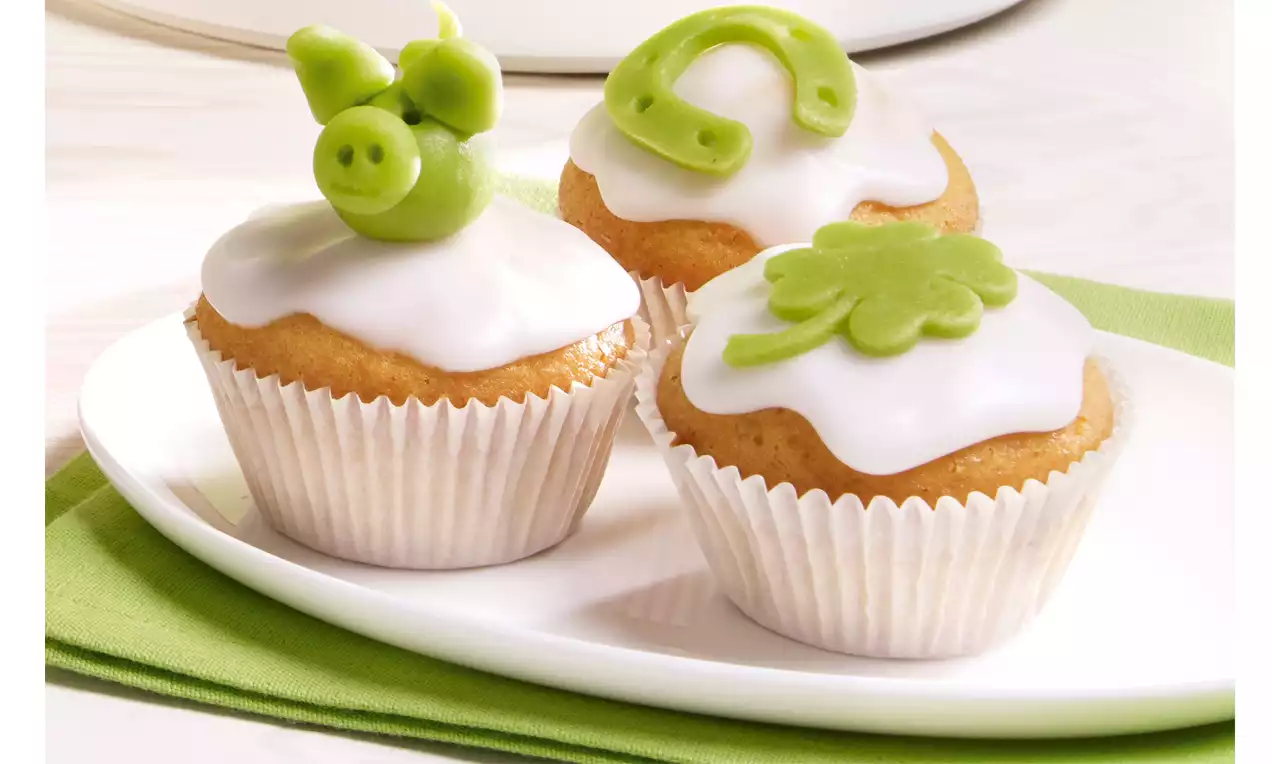 Drei goldbraune Muffins mit weißem Zuckerguss, dekoriert mit grünen Marzipanelementen wie Kleeblatt, Schweinchen und Hufeisen auf weißem Teller