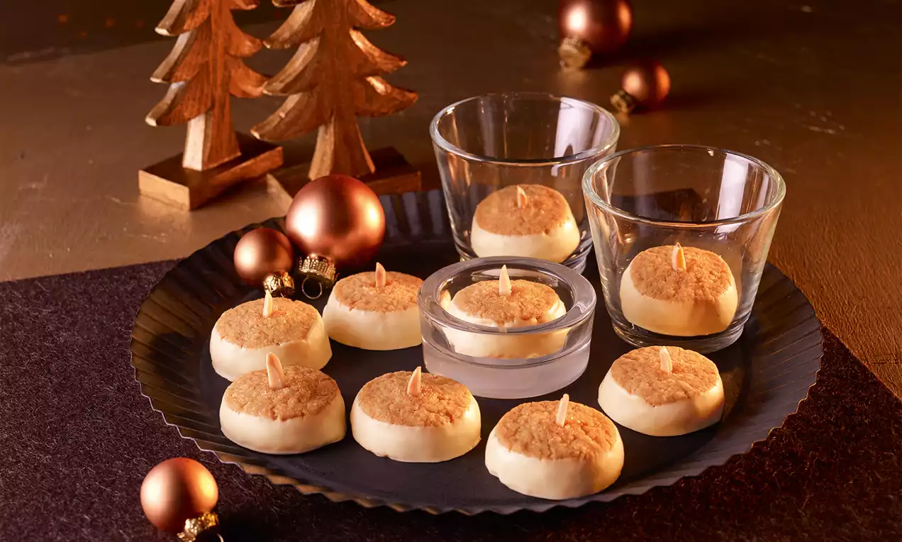 Yuvarlak, altın renkli tepsi üzerinde mum şeklinde tasarlanmış beyaz kurabiyeler, ahşap çam ağaçları ve bronz yılbaşı süsleri ile çevrili