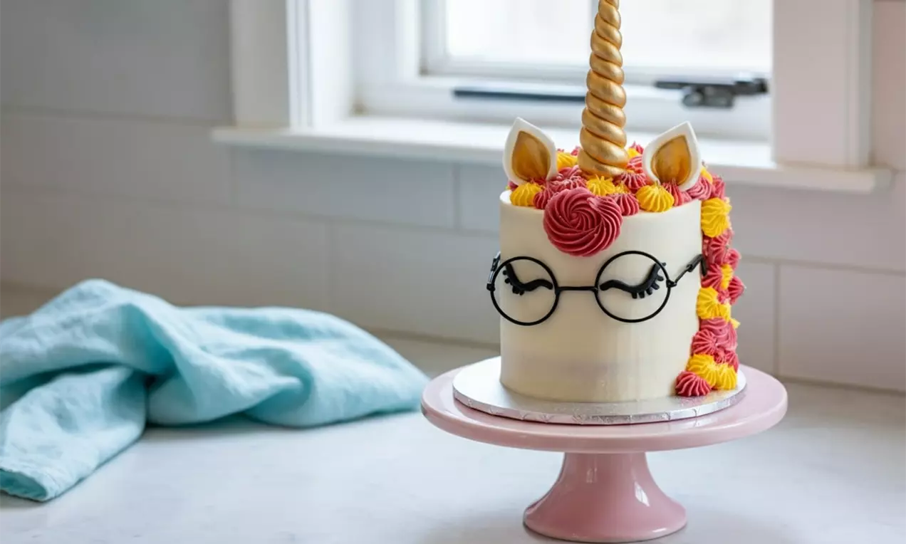 White cylindrical cake with golden horn, pink and yellow swirled mane, pointed ears, and glasses drawn in black, on a pink cake stand