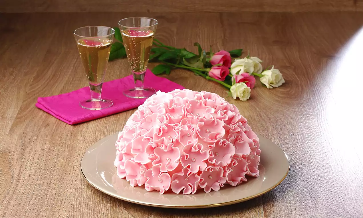 Pudra pembe ince kıvrımlı krema katmanlarıyla kaplı kubbe şeklinde pasta, açık renk bir tabakta, arka planda güller ve kadehlerle birlikte