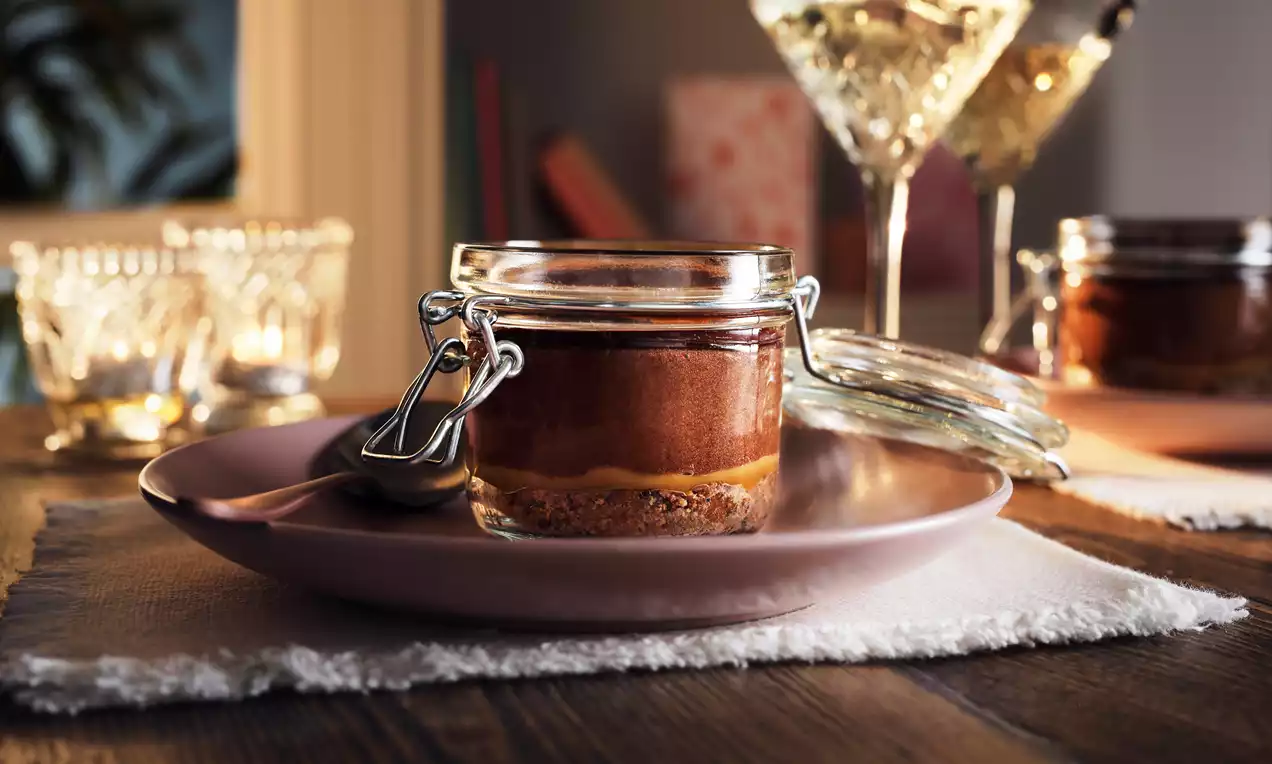 Chocolate Baileys mousse in a glass jar with layers of dark brown mousse, golden caramel, and crumbly biscuit base on a pink plate