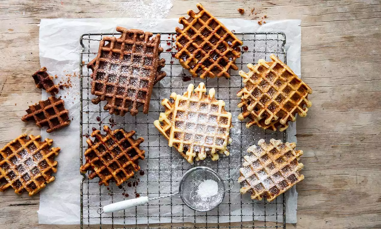 Waffle dorati e marroni disposti su una griglia, spolverati di zucchero a velo, con granelli di cioccolato su carta da forno e legno chiaro