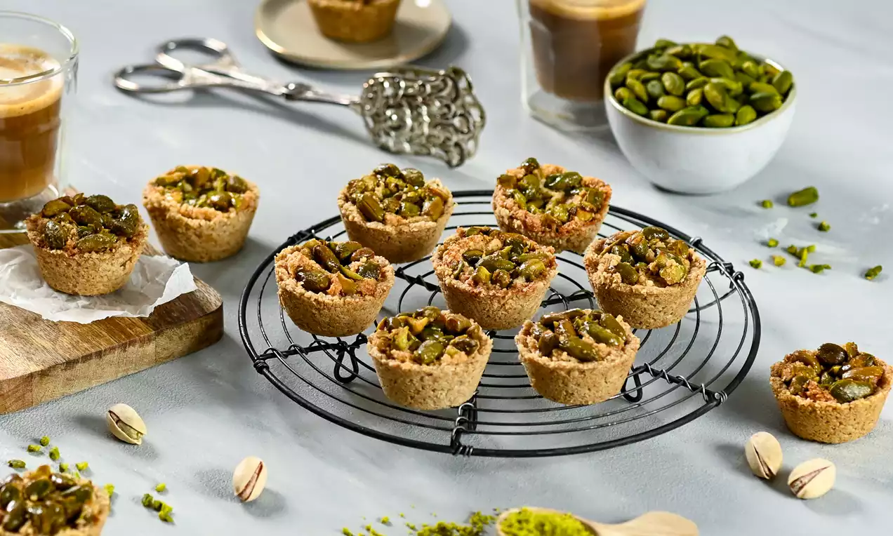 Gluten-free baklava cups with golden crumbly edges, topped with vibrant green pistachios, arranged on a black wire rack with coffee and pistachios nearby