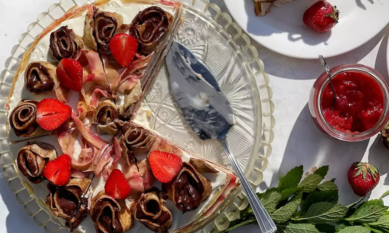 Palacinková torta s krémovou plnkou, vrstvami svetlého cesta, dekorovaná zrolovanými kakaovými palacinkami a čerstvými jahodami na sklenenom tanieri