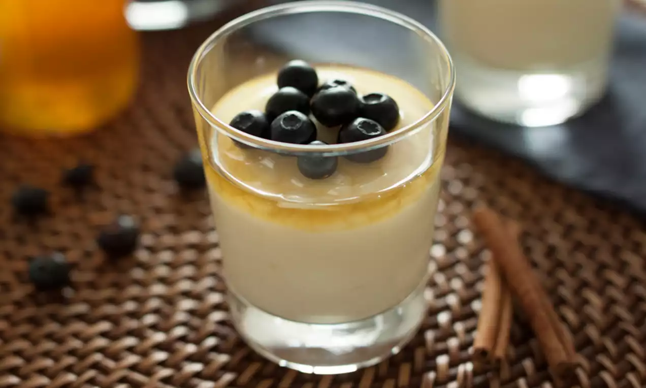 Crème vanillée lisse dans un verre transparent, nappée de sauce caramel dorée et garnie de myrtilles brillantes sur une table en osier