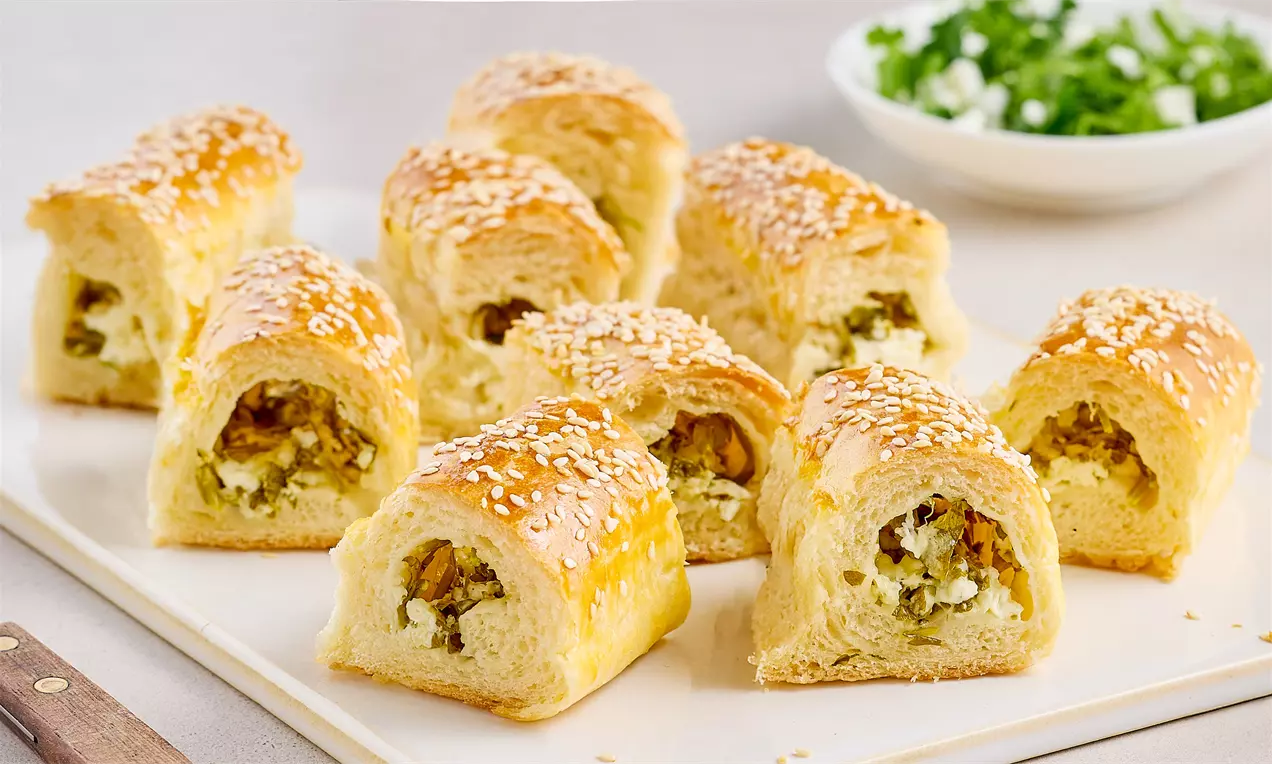 Altın rengi, susam serpilmiş peynirli tepsi ekmeği, iç dolgusu görünen kıtır üst yüzeyiyle dilimler halinde beyaz tabakta