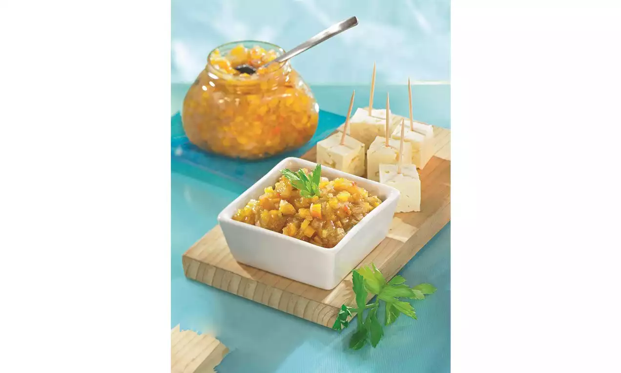 Broskyňové čatní žltooranžovej farby s jemnými kúskami v bielej keramickej miske, vedľa kocky syra na špajliach na drevenom podnose