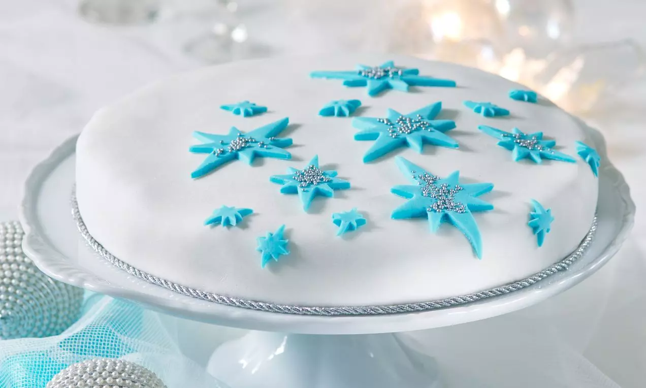 Vánoční dort s hladkým bielym poťahom, modrými ozdobami v tvare hviezd zdobenými striebornými glitrami, na bielom podnose s jemným lemom