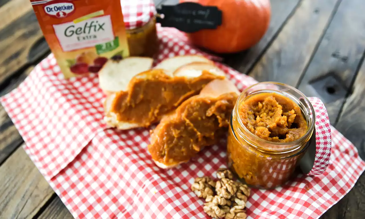 Tekvicový džem v sklenenom pohári s oranžovohnedou farbou, natretý na krajcoch chleba na červeno-bielej károvanej látke, vedľa orechov a tekvice