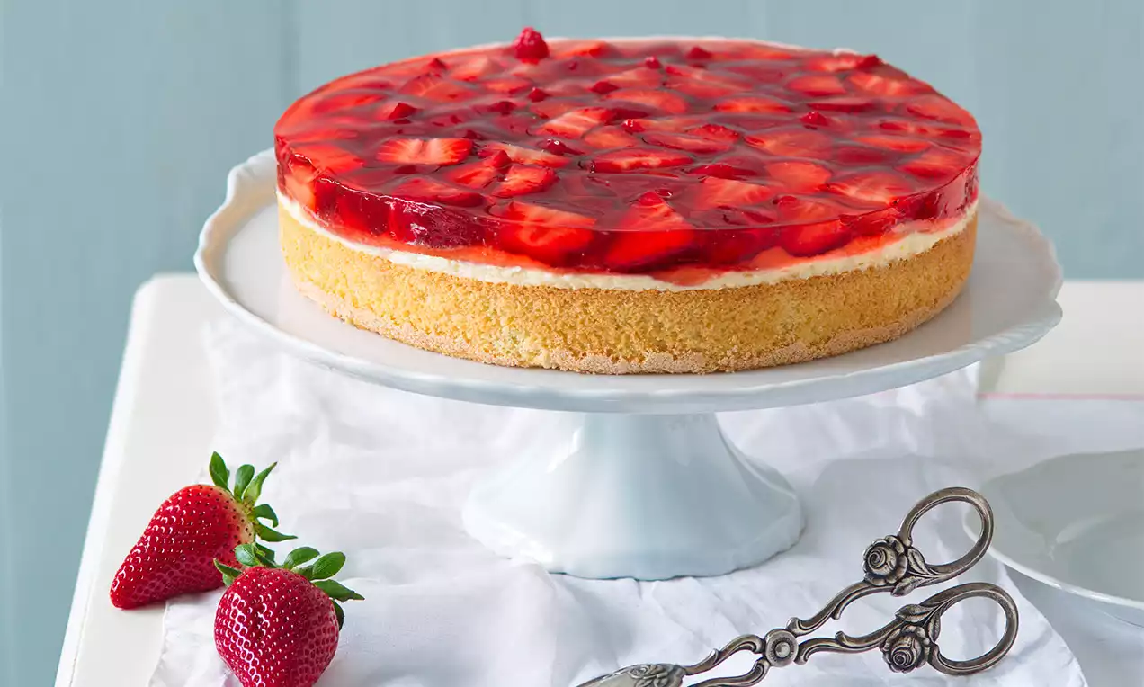Okrúhly ovocný dort na bielom podnose, so zlatistým piškótovým základom, krémovou vrstvou a lesklým želé s plátkami jahôd