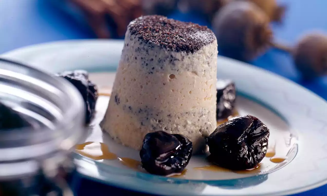 Mohn-Zimt-Parfait in zylindrischer Form mit heller, cremiger Textur, oben mit dunklem Mohn bestreut, umgeben von glänzenden Pflaumen und Sirup auf einem Teller