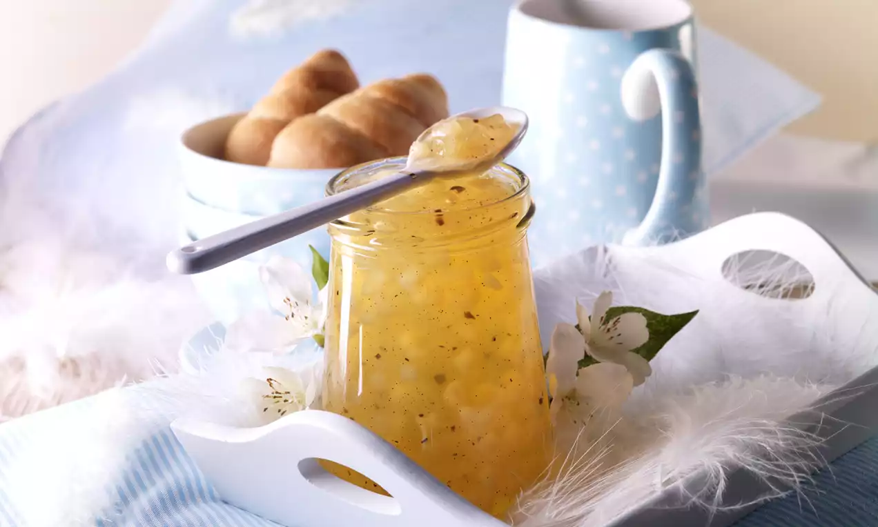 Žuti žele u prozirnoj staklenki s vidljivim komadićima kruške, ukrašen cvjetovima jabuke na bijeloj tacni i plavom pokrovu