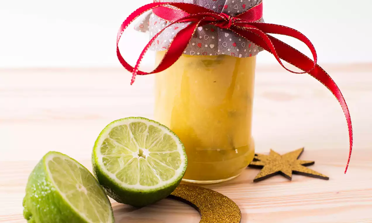 Sklenený pohár so žltým džemom, ozdobený sivým viečkom s červenou mašľou, vedľa polovíc limetky a trblietavých zlatých dekorácií
