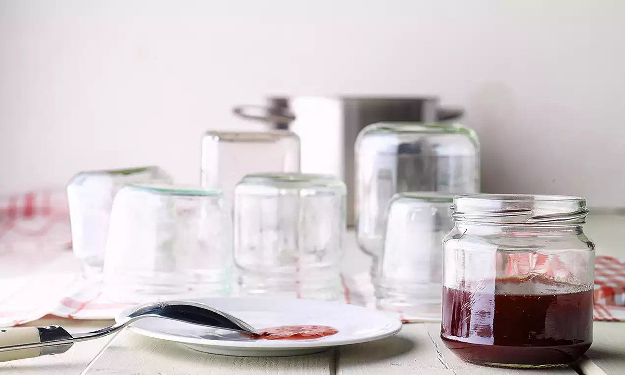 Campione di gelatina rosso scuro su un piatto bianco accanto a un cucchiaio, vasetti di vetro capovolti e una pentola sullo sfondo
