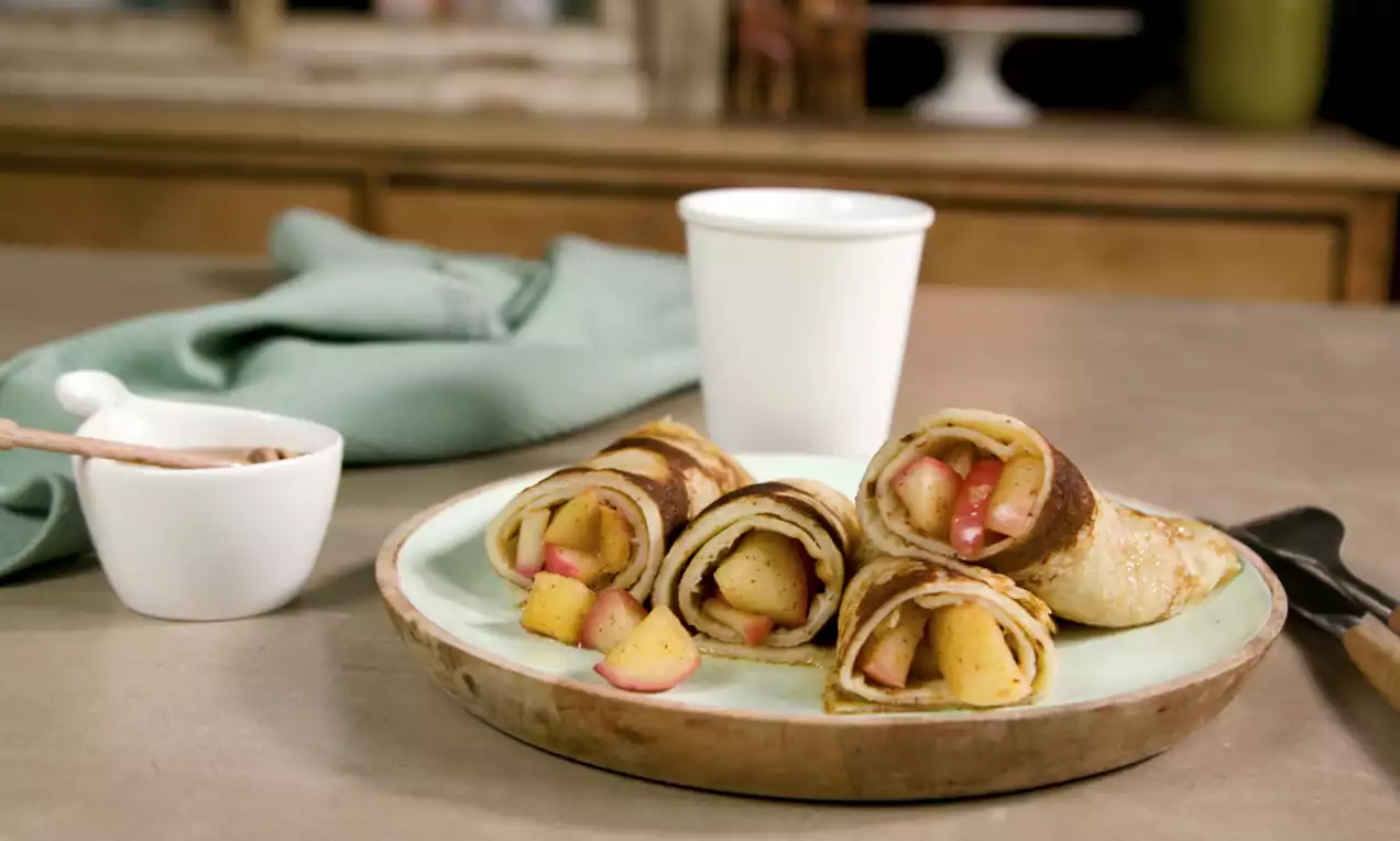 Opgerolde pannenkoeken met goudbruine randjes, gevuld met zachte kaneelappelstukjes op een lichtgroen bord met houten rand