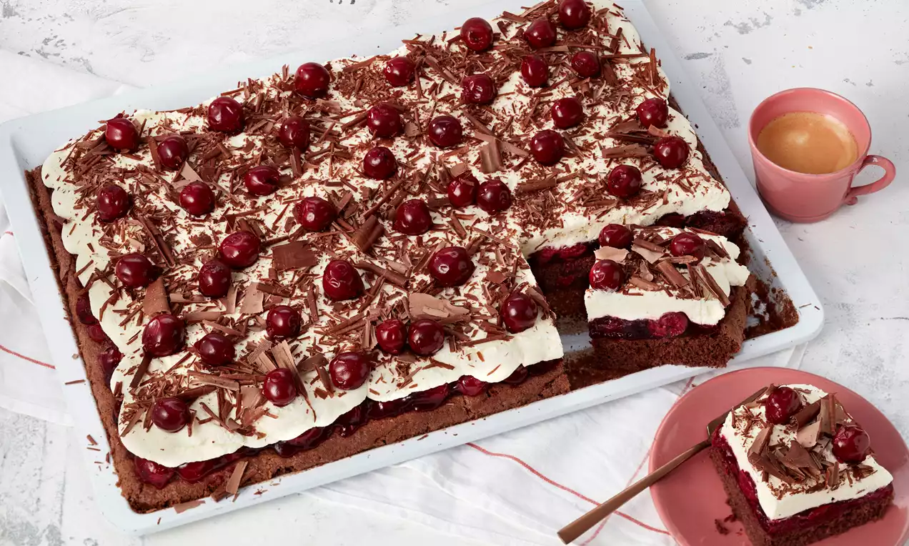 Schwarzwälder Kirschtorte vom Blech mit dunklem Boden, Sahneschicht, roten Kirschen und Schokoraspeln, daneben ein Stück auf rosa Teller