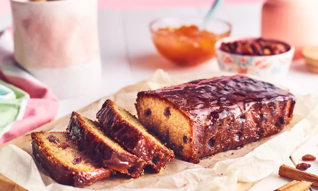 Golden-brown fruit and nut loaf with a glossy glazed top, studded with raisins, sliced on parchment paper with bowls and decor in the background