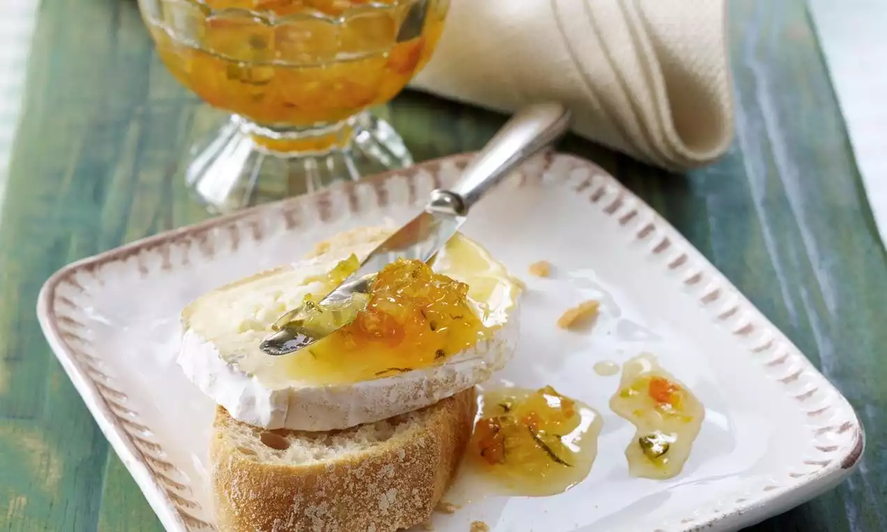 Gelb-orange Zucchinikonfitüre mit sichtbaren Kräutern auf Baguettescheiben und Käse, daneben etwas Konfitüre auf weißem Teller