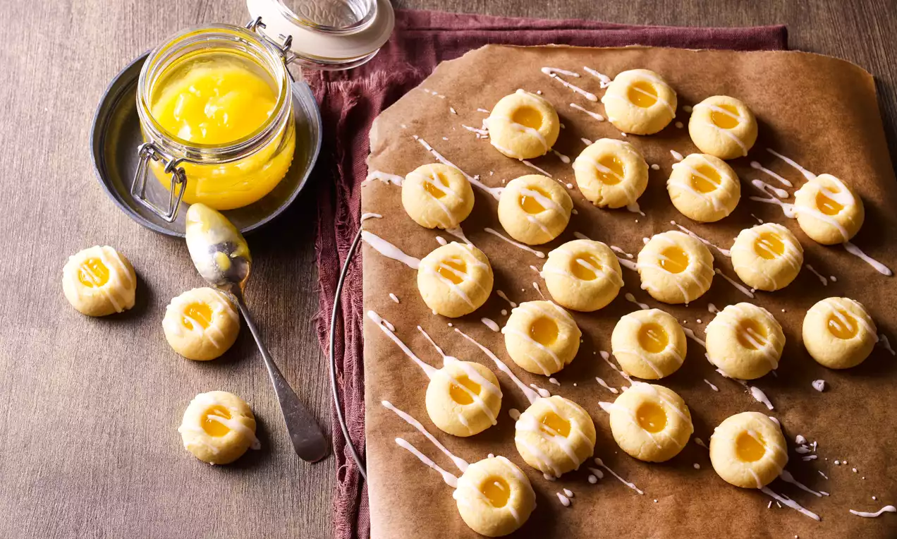 Sitni žućkasti keksići okruglog oblika s udubljenjem ispunjenim limun kremom, poslagani na smeđi papir s bijelim preljevima