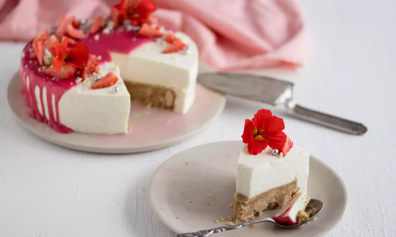 Hvid koldskaalskage med brun kiksebund og pink glaze, pyntet med røde blomster og jordbærstykker på lyse tallerkener