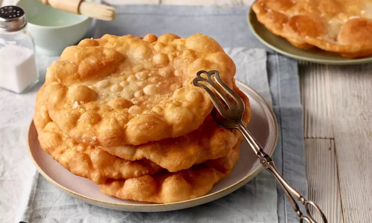 Goldgelber Langos, flach und leicht gewölbt mit knuspriger Oberfläche, gestapelt auf einem Teller, daneben eine silberne Kuchengabel