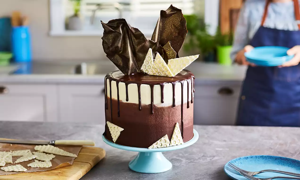 Chocolate ombre cake with smooth dark-to-light gradient, glossy chocolate drip, gold sails, white textured shards on a blue pedestal