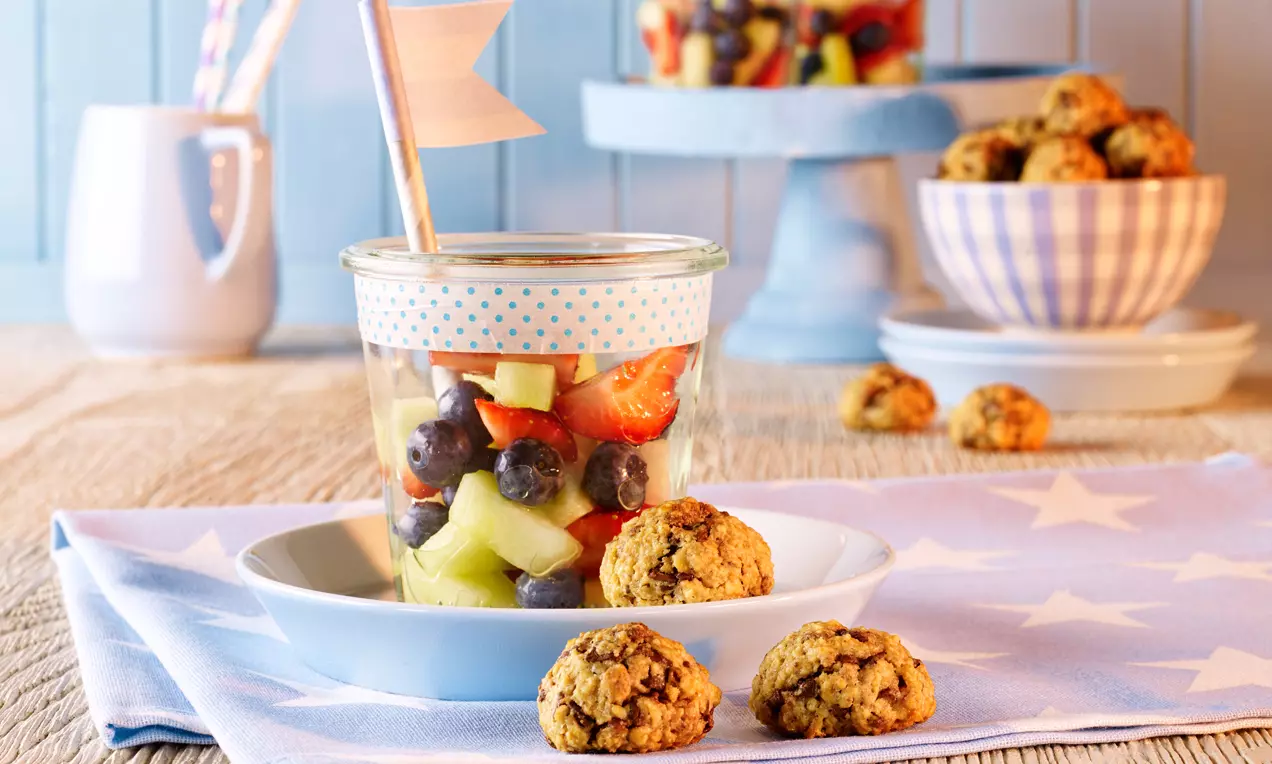 Bunte Erdbeer-Melonensalatwürfel mit Heidelbeeren in Glas mit Musterdeckel serviert, dekoriert mit Müslihappen auf pastelligem Stoff