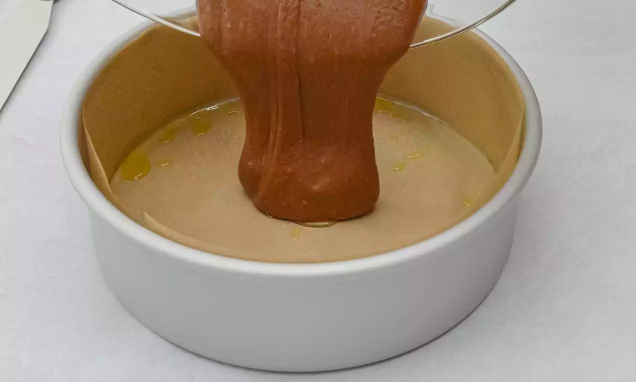 Chocolate batter flowing smoothly into a round white cake tin lined with light beige parchment on a pale surface