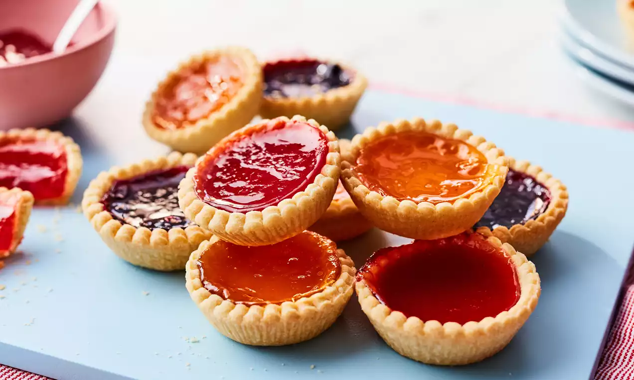 Golden jam tarts with crimped edges, glossy red, orange and purple fillings arranged on a pale blue board with a pink bowl nearby