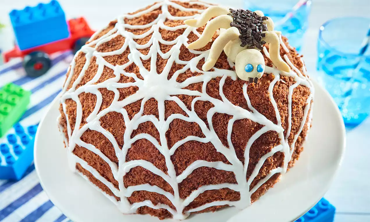 Okrúhla čokoládová torta s hnedým strúhovaným povrchom, pokrytá bielou cukrovou sieťou, ozdobená pavúkom z cesta s modrými očami na bielom tanieri