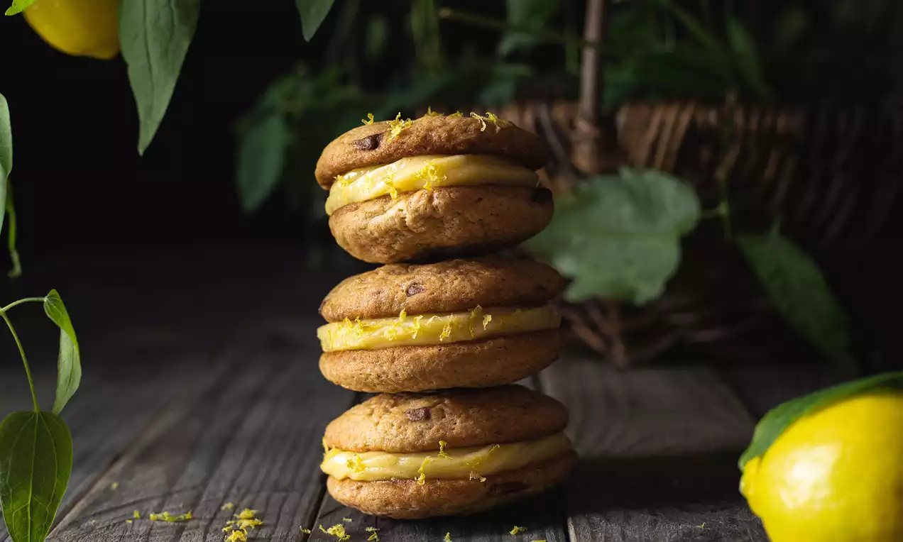 Tři zlatohnědé citronové cookies s jemnou texturou spojené vanilkovým krémem, posypané citronovou kůrou na dřevěném podkladu s citrony a zelenými listy