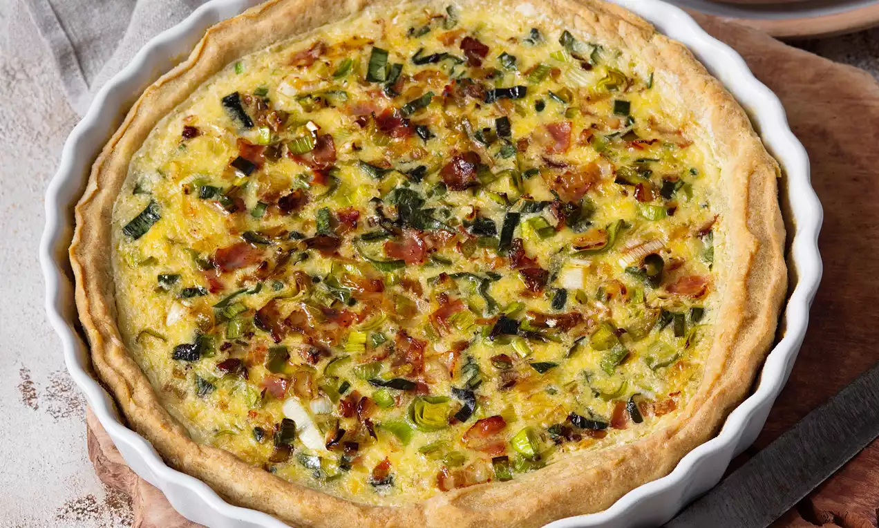 Pórkový koláč se zlatavou křupavou okrajovou krustou, pestrou náplní z pórku a slaniny, servírovaný v bílé vroubkované keramické formě