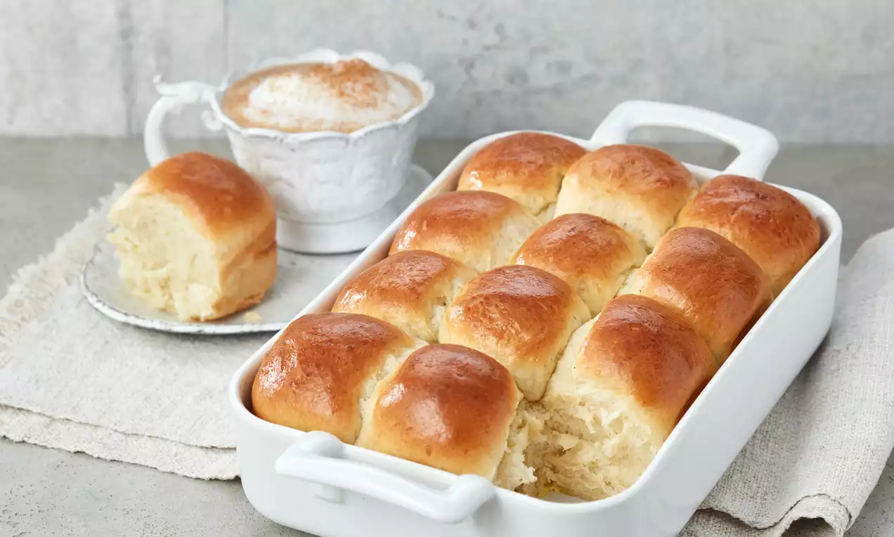 Goldbraune, glänzende Milchbrötchen in einer weißen Keramikform, ergänzt durch ein einzelnes Stück auf einem Teller neben einer Tasse Milchkaffee