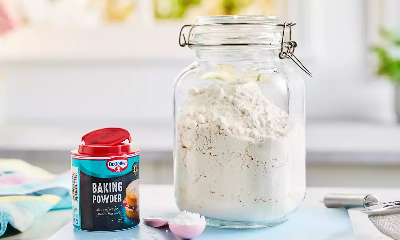 A clear jar filled with white flour on a light blue surface, next to an open red-lidded baking powder tin, a spoon, and pastel towels