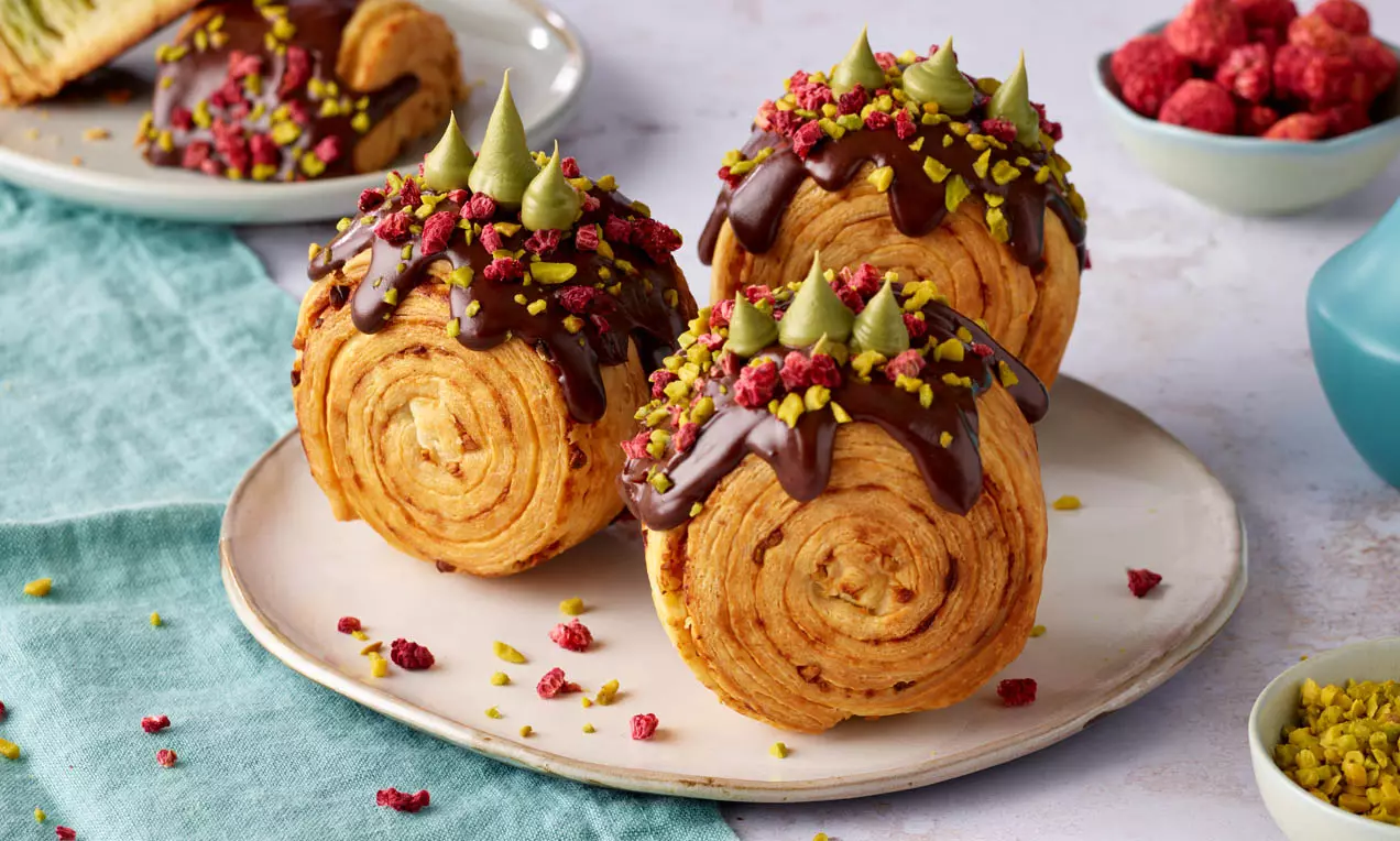 Goldene, spiralförmige Croissantrollen mit dunkler Schokoglasur, Pistazienstückchen, gefriergetrockneten Himbeeren und grünen Cremetupfen auf Keramikteller