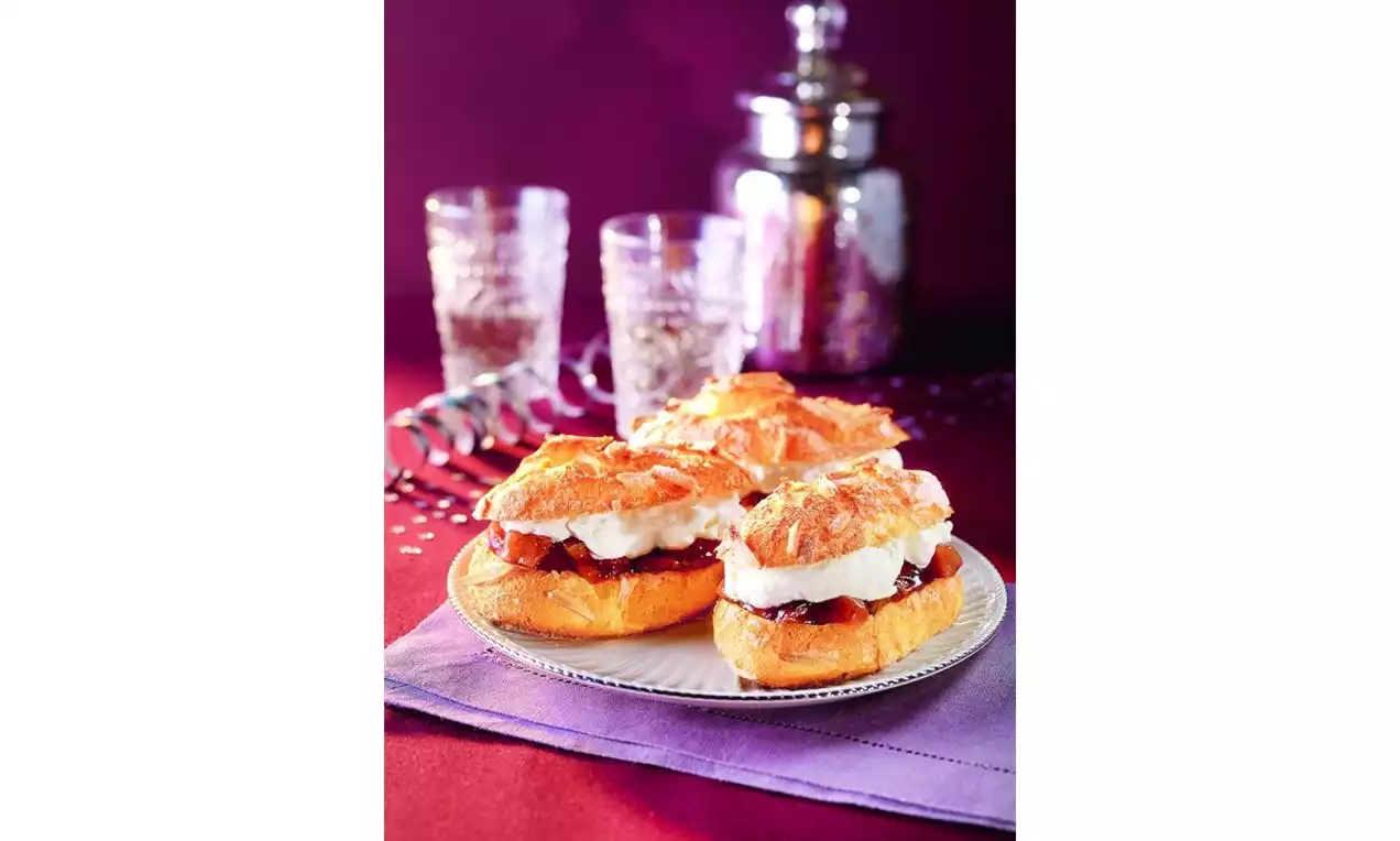 Zlatavé podlouhlé banánky s křupavým povrchem, plněné tmavou švestkovou náplní, šlehačkou a posypané skořicí na bílém talíři na fialovém ubrousku