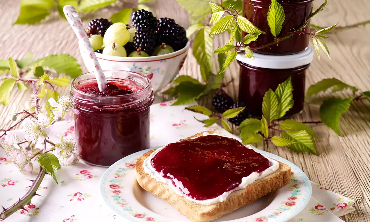Samtige Brombeer-Stachelbeer-Konfitüre im Glas, tiefviolett glänzend, mit Brombeeren, Stachelbeeren und einer Zimtstange auf Holzbrett