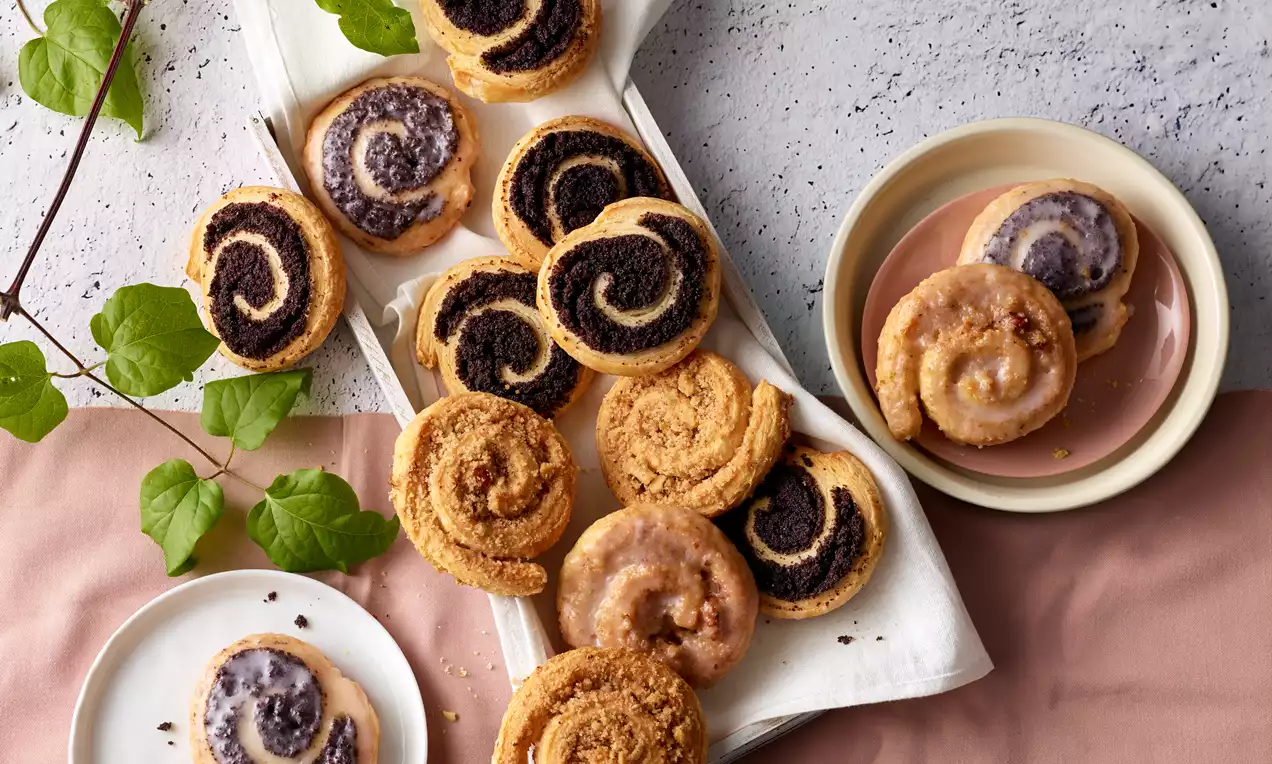 Kleine Schnecken aus hellem Teig mit dunkler Mohnfüllung und goldbraunen Nussstücken auf Tellern und weißem Tuch, dekoriert mit grünen Zweigen