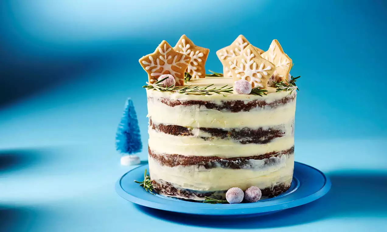Layered gingerbread cake with creamy frosting, topped with snowflake cookies, rosemary sprigs, and sugared berries on a blue plate
