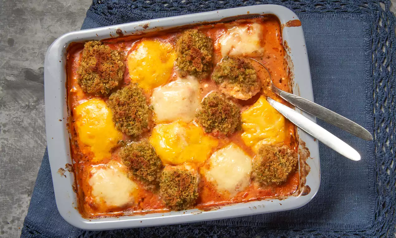 Goldbraune Hähnchenschnitzel mit Kräuterkruste, schmelzendem Käse und roter Tomatensoße in einer grauen Auflaufform auf dunkelblauer Stoffunterlage