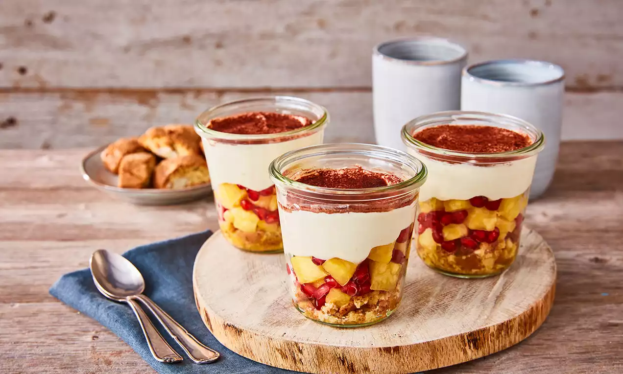 Schichtdessert in Gläsern mit hellgelbem Ananasstückchen, rubinrotem Granatapfel, cremefarbener Mascarponecreme und braunem Kakaopulver