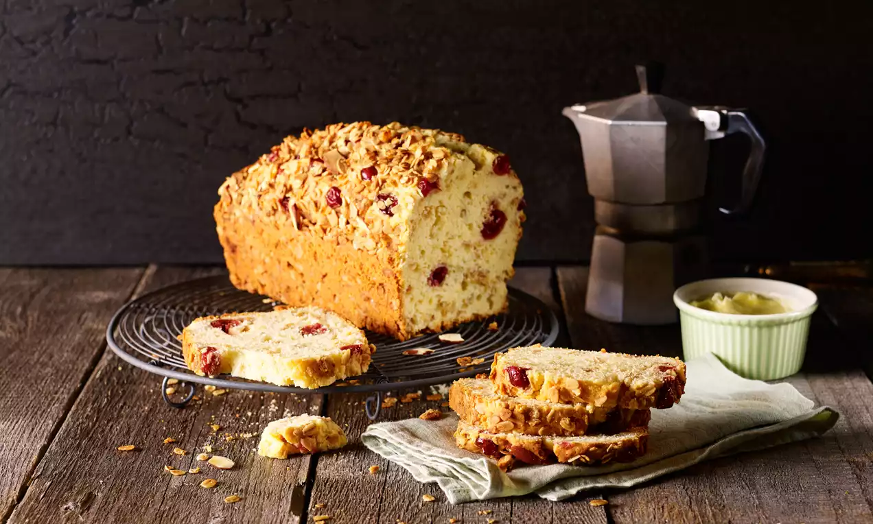 Müsli-Cranberry-Kuchen mit goldbrauner Kruste und roten Cranberry-Stücken auf einem Rost, neben geschnittenen Scheiben auf einem Tuch