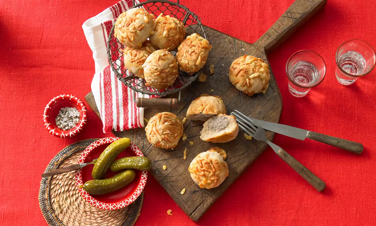 Okrugli zalogajčići od kruha zlatnosmeđe korice posuti sirom, složeni na drvenoj dasci uz vilicu, krastavce i crvenu dekoraciju