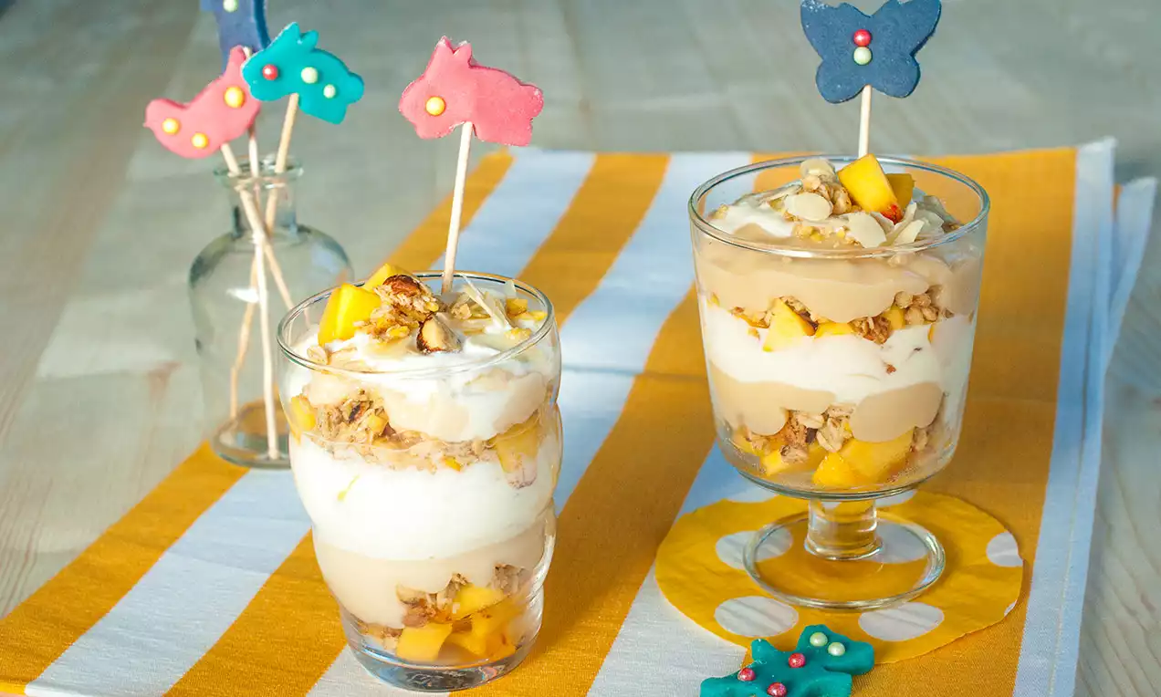 Vrstvený mandlový a broskvový dezert ve sklenici se střídajícími se krémovými vrstvami, kousky broskví a posypanými ořechy, dekorovaný barevnými sušenkami