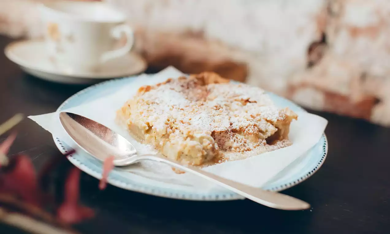 Raparperipiirakka vaaleanmuruisella pinnalla, tomusokerilla kevyesti peitettynä, aseteltuna siniselle lautaselle lusikan kanssa