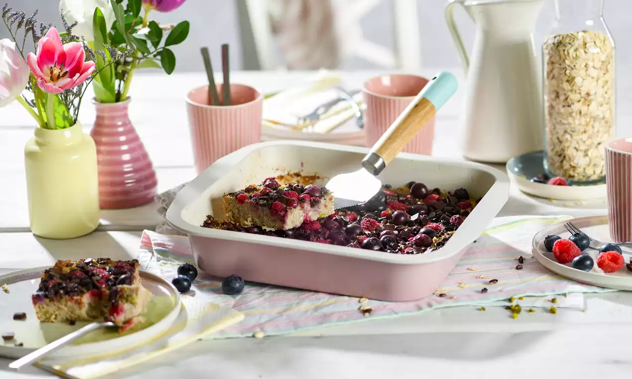 Haferflockenkuchen mit braunem Boden und saftiger Beerenfüllung in rosafarbener Backform, arrangiert mit Obst und Geschirr auf hellem Tisch