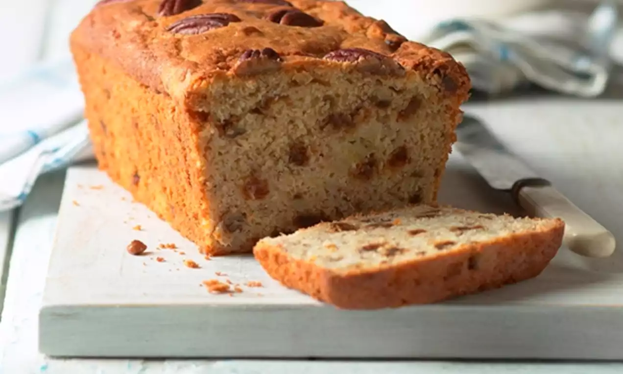 Golden-brown loaf cake studded with pecans and raisins on a white wooden board, one slice cut and lying flat beside a knife