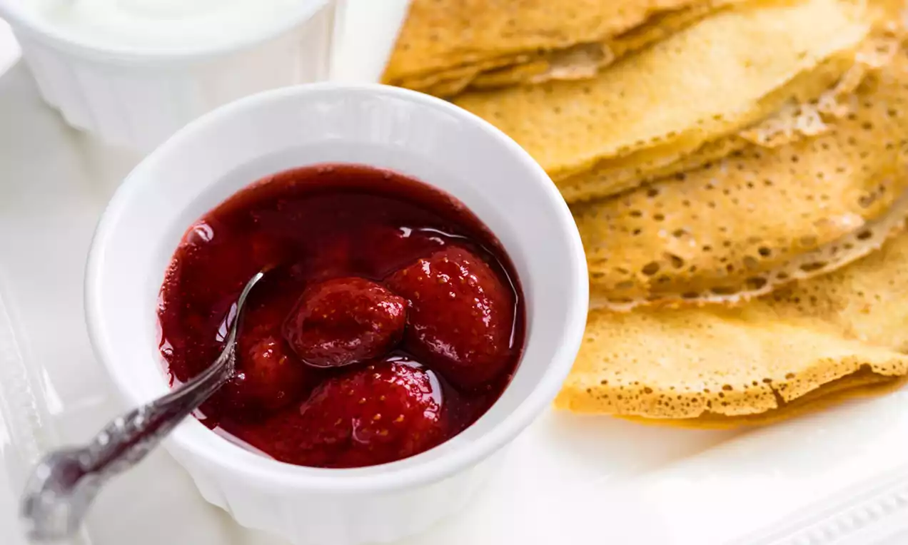 Gem de căpșuni roșu-intens cu textură lucioasă și bucăți întregi, servit într-un bol alb alături de clătite aurii și smântână cremoasă