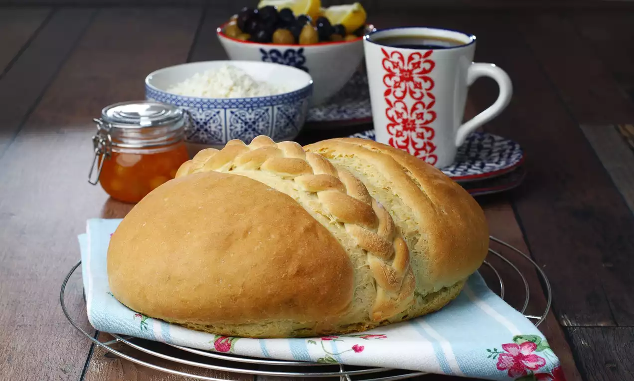 Altın sarısı yuvarlak Trabzon ekmeği, örgü süslemeli kabuğu ile desenli bir örtü üzerinde tel ızgarada, kahvaltılıklarla sunulmuş
